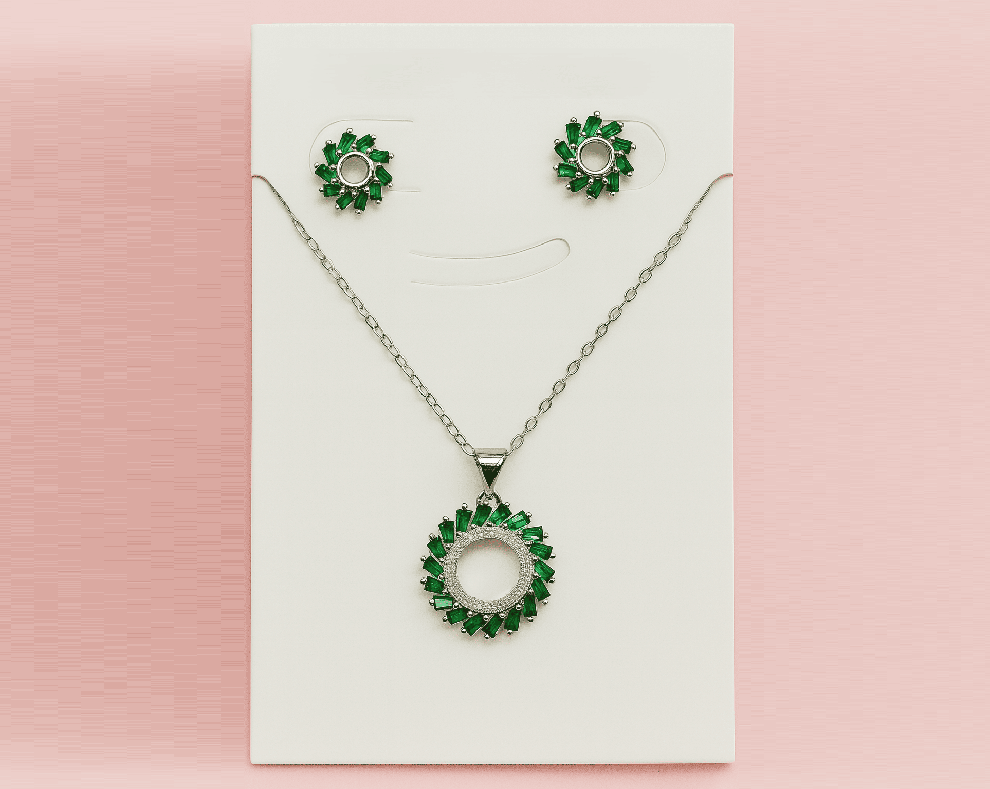 Elegant green halo pendant necklace and stud earrings set with emerald green stones displayed on white board against pink background