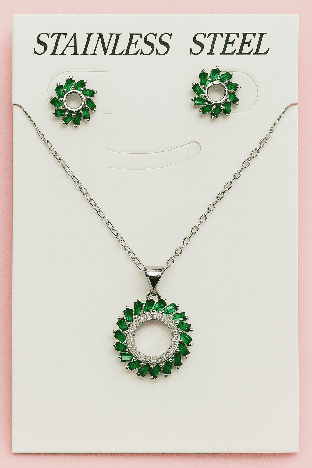 Green halo pendant necklace and stud earrings set with emerald green stones displayed on white card labeled 'Stainless Steel' against pink background
