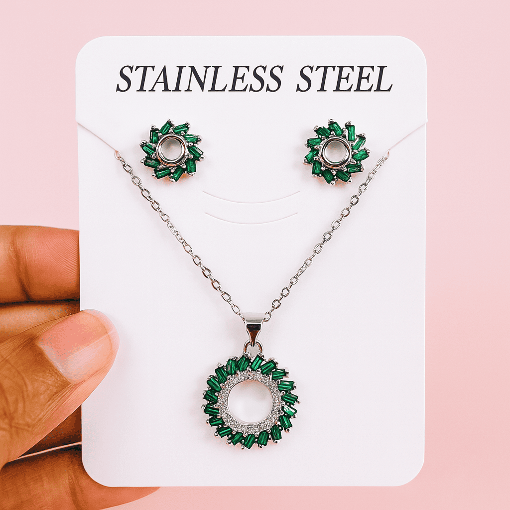 Emerald green halo pendant necklace and stud earrings displayed on a white 'Stainless Steel' card held by hand against pink background