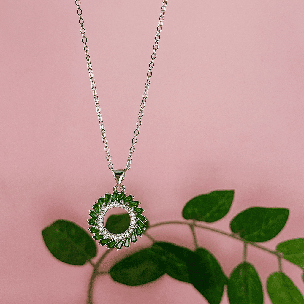 Silver halo pendant necklace with emerald green stones hanging above green leaves against pink background