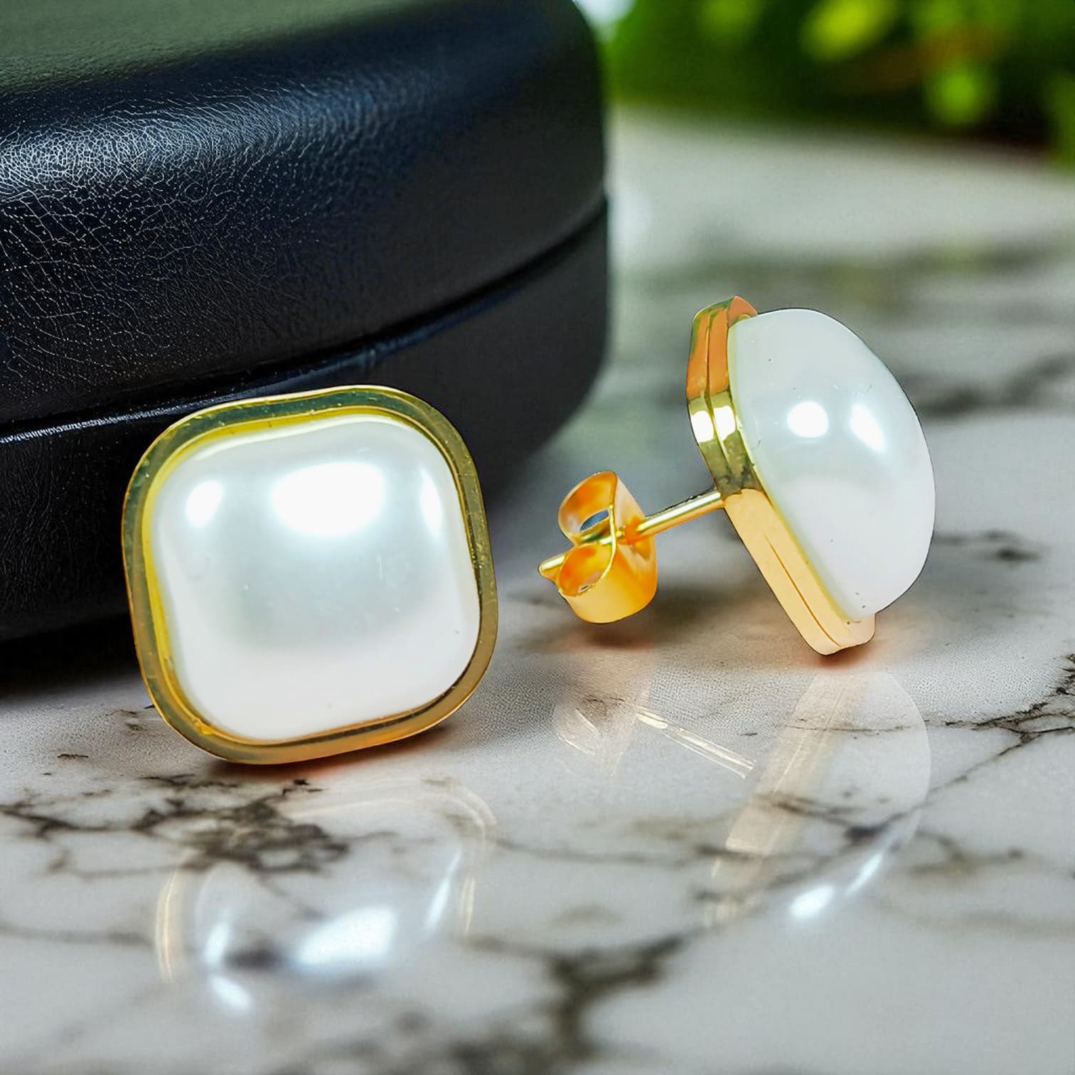 Square pearl stud earrings with gold-plated trim displayed on marble surface near black jewelry box