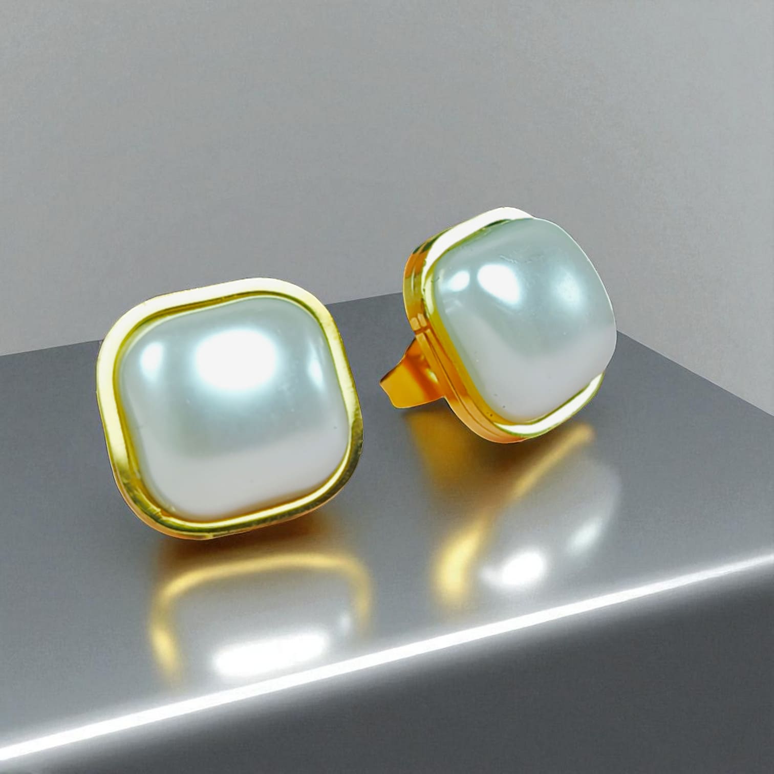 Square pearl stud earrings with gold-plated frames on reflective metallic surface