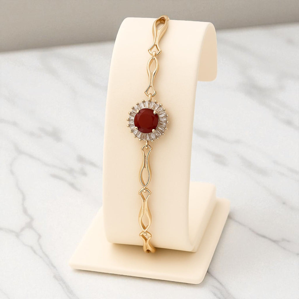Gold-tone bracelet with red gemstone and zircon accents on beige stand against marble background